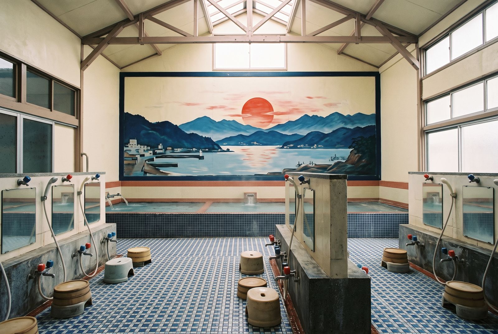 ミナトの浴室。正面の壁に港と山と夕日を描いた大きなペンキ絵があり、手前に青いタイルの湯船と洗い場が並ぶ