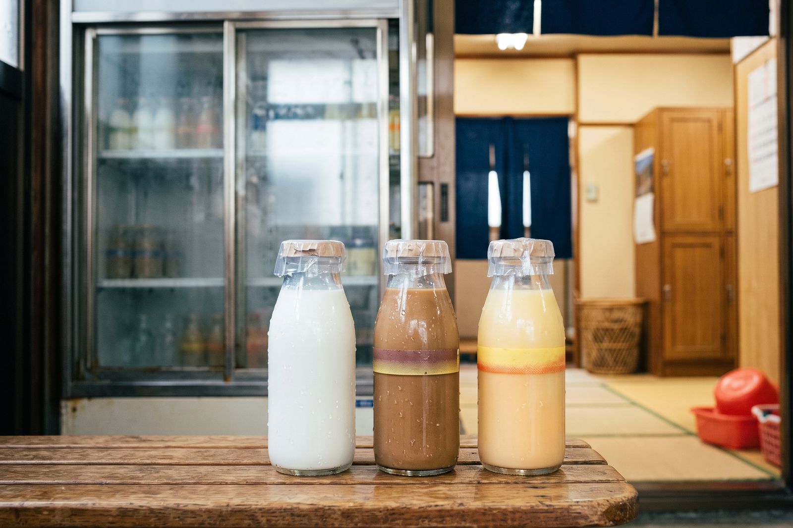 木の台に並んだ冷えた瓶の牛乳、コーヒー牛乳、フルーツ牛乳。奥に脱衣所が見える